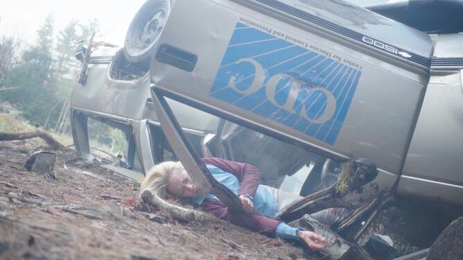 Kristanna Loken lying injured and trapped under an overturned vehicle in a forest setting, portraying the character Alex in the survival thriller 'DARK NIGHT OF THE SOUL.'