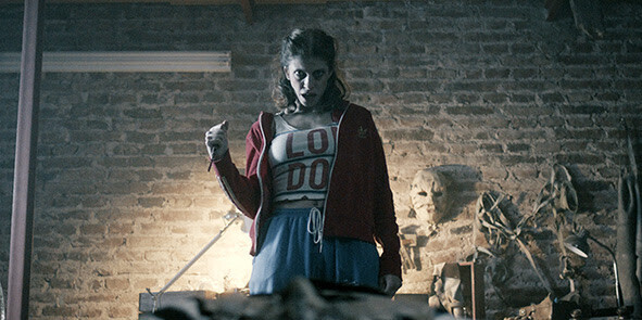 A tense scene from 'PLAY DEAD' shows a young woman standing in a dimly lit basement with an intense, determined expression. She wears a red zip-up hoodie over a crop top with bold letters and blue shorts. Her clenched fist and piercing gaze suggest defiance or confrontation. Behind her, a brick wall and eerie, shadowy objects—including a sculpted face—add to the unsettling atmosphere. The lighting casts dramatic shadows, heightening the suspense and horror elements of the image.