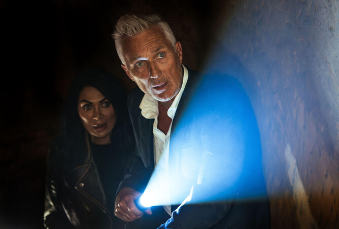 Martin Kemp as Detective Verney and Jeanine Nerissa Sothcott as Francesca shine a torch in a darkened space, searching for clues in 'DOCTOR PLAGUE.'