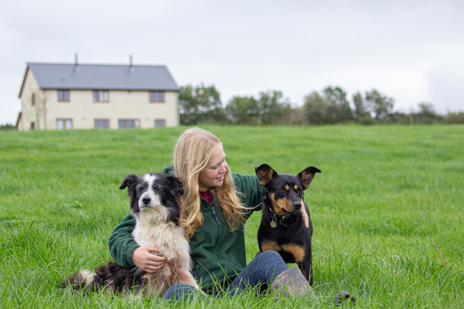 Amy smiling, while sitting with her arms round Mist the Border Collie & Izzy the Kelpie in the grassland in front of the house on the horizon