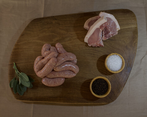 wooden board from above displaying a pile of sausages and three chops with bowls of rock salt and peppercorns & a sprig of sage